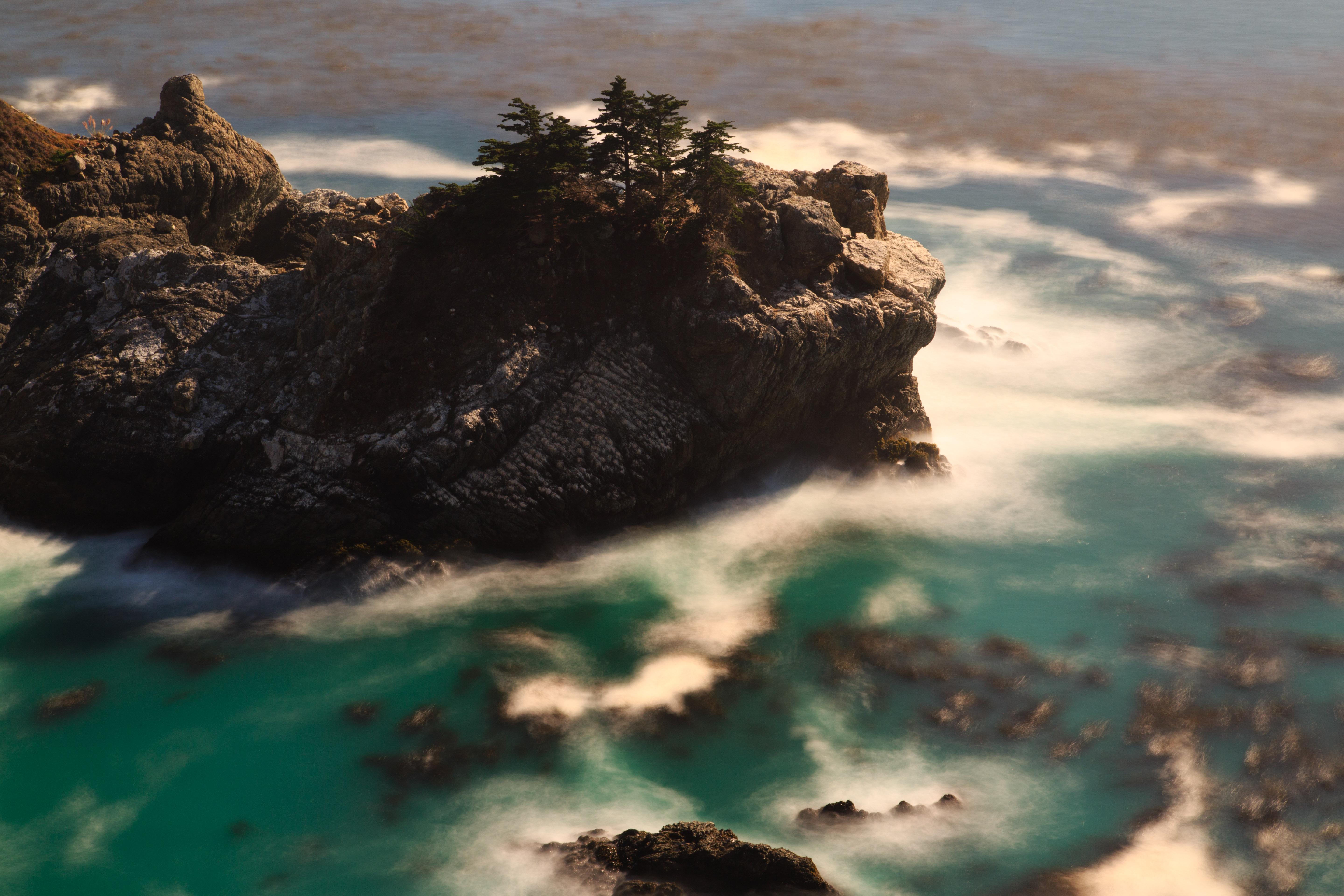 Rocky coastline with trees perched atop a sea stack, surrounded by turquoise water